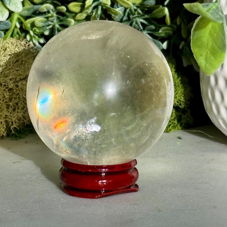Op de afbeelding: Een heldere kristallen bol met interne insluitsels, rustend op een glanzende, donkerrode houten standaard. De bol heeft een lichte gele tint en vertoont een regenboogeffect door lichtbreking. De achtergrond bevat groen en een wit decoratief element.
