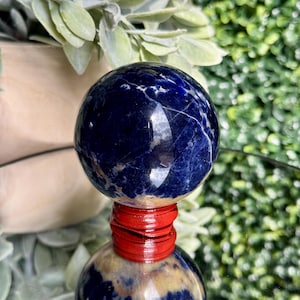 May include: A polished, dark blue sodalite sphere with white veining, resting on a red wooden base. The sphere is displayed on a reflective surface, with greenery in the background. A decorative crystal for home decor.