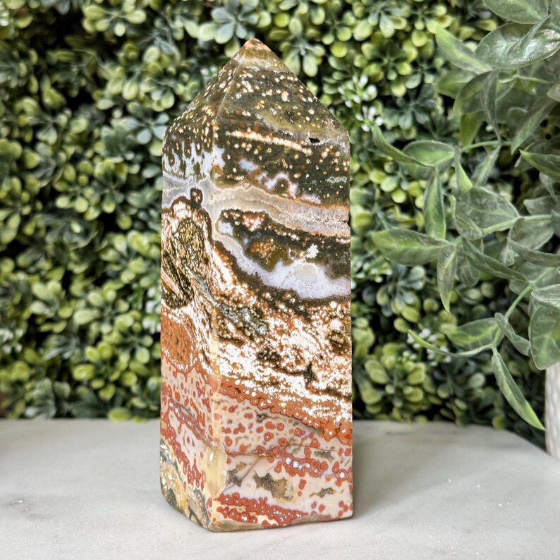 May include: A polished stone obelisk with a pointed top. The stone features a mix of colors, including white, brown, red, and green, with a speckled and layered pattern. The obelisk is a decorative item.