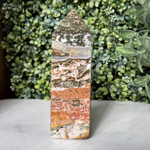May include: A polished, rectangular gemstone tower with a pointed top. The stone features a mix of colors, including white, brown, orange, and green, with a speckled and layered pattern. The tower is standing upright.