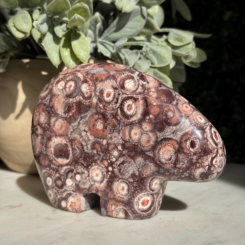 May include: A carved stone sculpture in the shape of a bear. The sculpture is a mix of brown, red, and cream colors with a unique pattern of circular designs. The bear has a smooth, polished surface and is displayed in natural light.