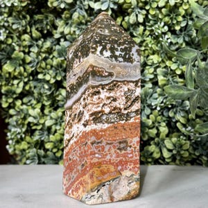 May include: A polished, rectangular ocean jasper obelisk. The stone features a mix of colors, including green, brown, white, and red, with a speckled pattern. The obelisk has a pointed top and is displayed against a backdrop of green foliage.