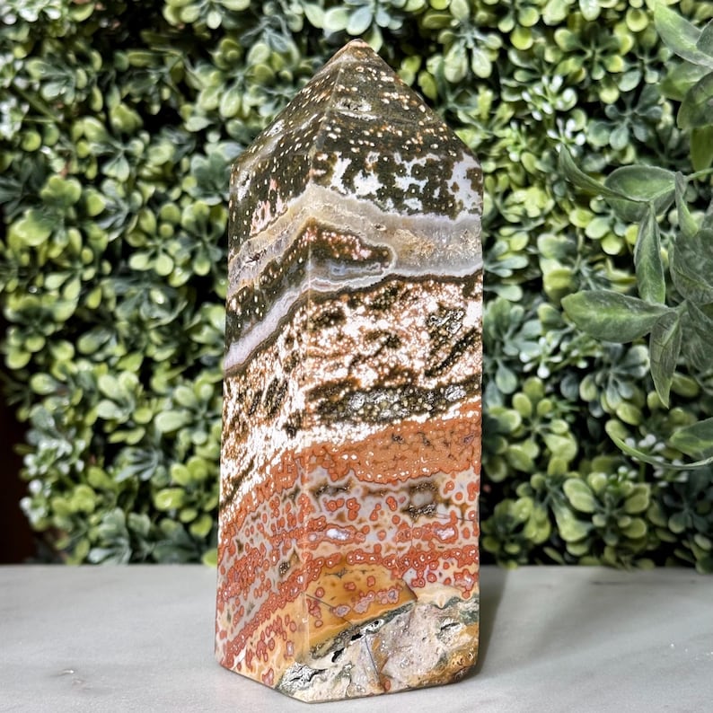 May include: A polished, rectangular ocean jasper obelisk. The stone features a mix of colors, including green, brown, white, and red, with a speckled pattern. The obelisk has a pointed top and is displayed against a backdrop of green foliage.