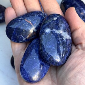 May include: Several polished sodalite stones in varying sizes. The stones are a deep blue color with white and gray veining. The stones are smooth and have a glossy finish. The stones are held in a hand.