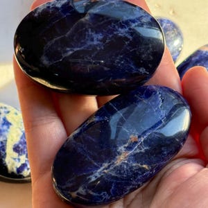 May include: Several polished sodalite stones in shades of deep blue with white marbling. The stones are oval-shaped and smooth, with a glossy finish. The stones are held in a hand.