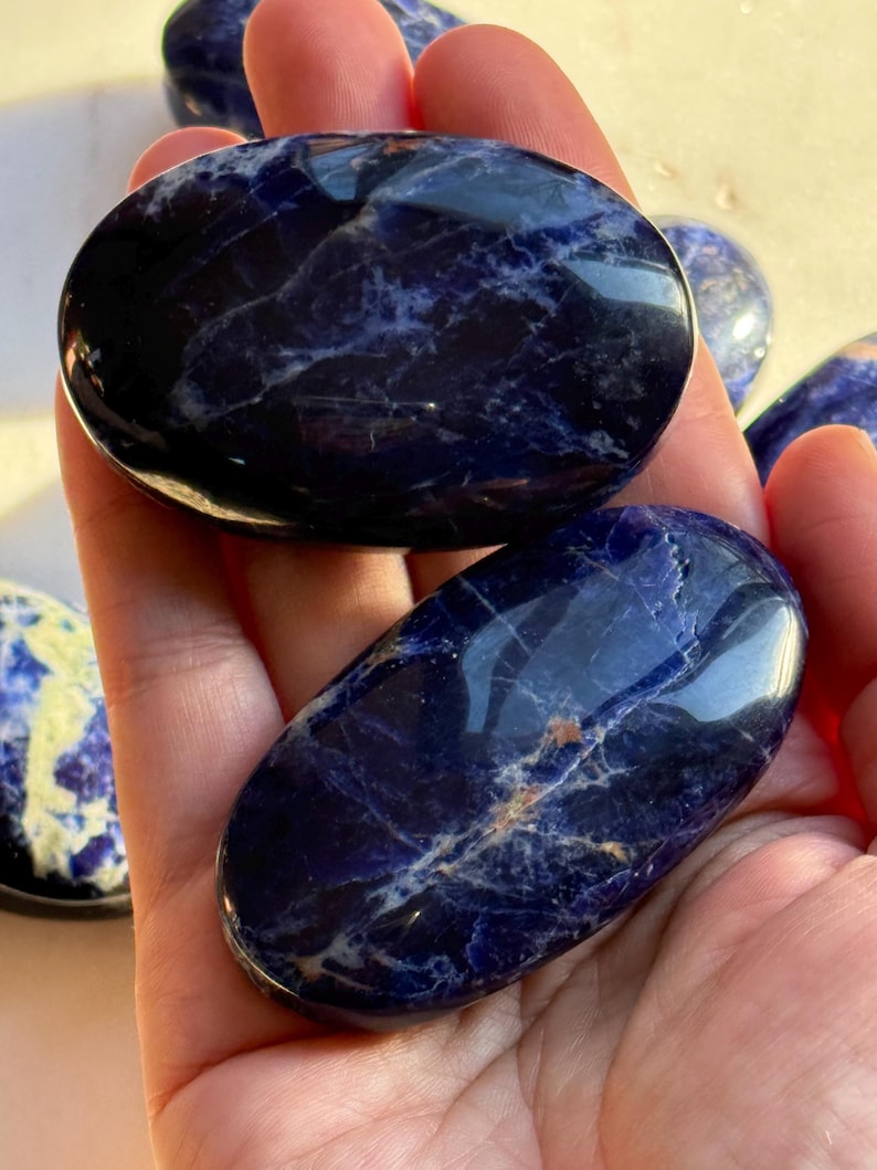 May include: Several polished sodalite stones in shades of deep blue with white marbling. The stones are oval-shaped and smooth, with a glossy finish. The stones are held in a hand.