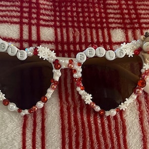 May include: Heart-shaped sunglasses with brown lenses, adorned with white beads, red accents, and the words "JINGLE BELLS." Features Santa Claus and reindeer figurines on the frames, perfect for Christmas.