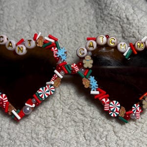 May include: Heart-shaped sunglasses with brown lenses, decorated with red, white, and green embellishments. The text "CAN'T CATCH ME" is spelled out in letter beads. Gingerbread men and candy cane accents add a festive touch.