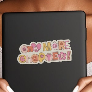 Puede incluir: Una pegatina con el texto "one more chapter!" en letras de colores, con contorno, con flores y un corazón. La pegatina está en un portátil negro.