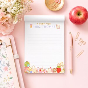 May include: A white notepad with horizontal lines, featuring the text A Note From MRS. THOMAS at the top. The bottom border is decorated with colourful illustrations of school supplies, flowers, and a coffee cup. A pink pen sits next to the notepad.
