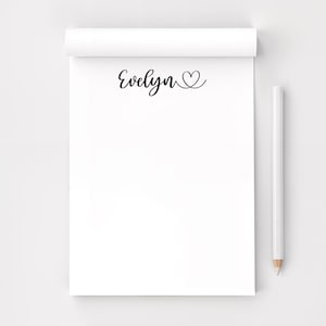 Peut inclure: Un bloc-notes blanc avec le nom "Evelyn" en écriture cursive et un symbole de cœur. Un crayon blanc est posé à côté du bloc-notes. Le bloc-notes est enroulé en haut.