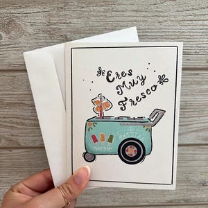 May include: Greeting card with a hand-drawn illustration of a turquoise ice cream cart with the words "Eres Muy Fresco". The cart has colorful popsicles and a sign. The card is held with a white envelope.