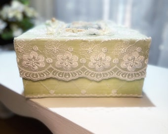 Puede incluir: Una funda para caja de pañuelos de color verde claro con un borde de encaje blanco. El encaje tiene un patrón floral.
