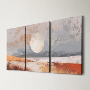 May include: Abstract landscape art print set. The artwork features a large, pale circle in the center panel, resembling a sun or moon. The color palette includes shades of gray, orange, and white, creating a serene and minimalist aesthetic. The set is composed of three vertical panels.