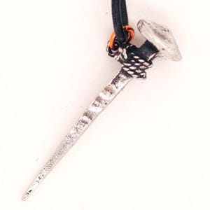 May include: A silver-coloured nail pendant with a textured finish hangs from a black cord. The nail head is flat and round, and the nail shaft is long and tapered. An orange ring connects the cord to the pendant.