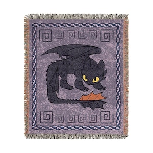 May include: A woven throw blanket featuring a black dragon with yellow eyes and orange accents. The dragon is centered on a lavender background with a repeating geometric border and fringe.