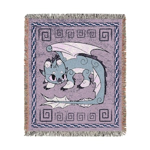 May include: A woven throw blanket featuring a cartoon dragon in shades of blue, white, and lavender. The dragon has large wings and is set against a lavender background with a geometric border and fringe.