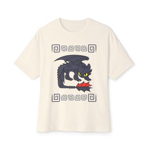 May include: Cream-colored t-shirt featuring a cartoon dragon with black and red accents. The dragon is surrounded by a decorative square border. The shirt has short sleeves and a crew neck.