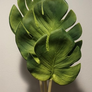 May include: A bunch of three artificial green monstera leaves with stems.