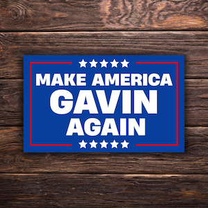 Può includere: Un cartello blu con la scritta bianca "MAKE AMERICA GAVIN AGAIN", incorniciato da un bordo rosso. Il cartello ha cinque stelle bianche in alto e in basso, su uno sfondo di legno.