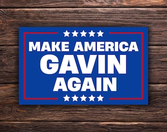 Può includere: Un cartello blu con la scritta bianca "MAKE AMERICA GAVIN AGAIN", incorniciato da un bordo rosso. Il cartello ha cinque stelle bianche in alto e in basso, su uno sfondo di legno.