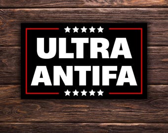 Può includere: Un adesivo rettangolare nero con la scritta "ULTRA ANTIFA" in grassetto bianco. Il design include un bordo rosso e sei stelle bianche sopra e sotto il testo. Lo sfondo è un legno scuro.