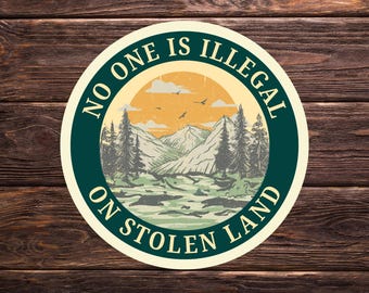 Può includere: Un adesivo rotondo con un paesaggio di montagne, alberi, un tramonto e uccelli. Il testo "NO ONE IS ILLEGAL ON STOLEN LAND" è sul bordo verde scuro, su uno sfondo color crema.