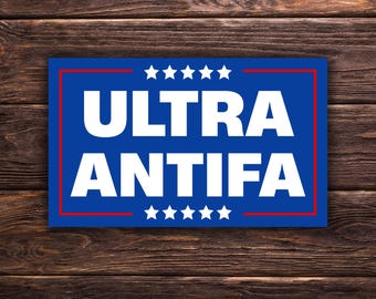 Può includere: Un cartello rettangolare blu con le parole "ULTRA ANTIFA" in grandi lettere maiuscole bianche. Il cartello ha un bordo rosso e sei stelle bianche sopra e sotto il testo. Lo sfondo è una superficie di legno marrone scuro.