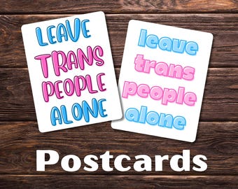 Può includere: Due cartoline bianche con la scritta "LEAVE TRANS PEOPLE ALONE" in lettere blu e rosa. La parola "Postcards" è scritta in bianco in basso nell'immagine. Le cartoline sono su una superficie di legno scuro.