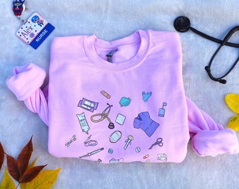May include: Pink sweatshirt with a design of various medical tools and the word "NURSE" on a name tag. A stethoscope and fall leaves are also in the image.