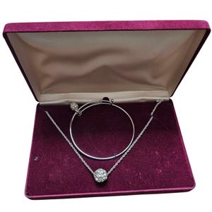 Può includere: Un bracciale rigido in metallo color argento e una collana a catena ritorta abbinata, entrambi dotati di un ciondolo sferico incastonato con cristalli trasparenti. Il set di gioielli è presentato all'interno di un cofanetto rettangolare in velluto viola.