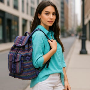 May include: A multi-colored backpack with a drawstring closure and two front pockets. The backpack features a woven pattern in shades of blue, red, and yellow. The person is wearing a turquoise button-down shirt and white shorts.