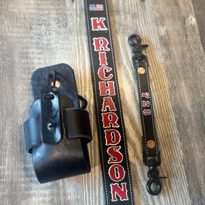 May include: A black leather radio holder, a black leather strap with the name "RICHARDSON" in red letters, and a black strap with the number "728" in red. The items are on a wooden surface.