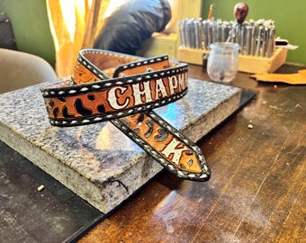 May include: A tan leather belt featuring a dark brown leopard print pattern and white stitching along the edges. The belt is embossed with the name CHAPMAN in white capital letters, with a pointed end and a buckle attachment point.