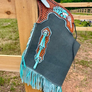 May include: A handcrafted leather chaps with a turquoise and brown design. The chaps feature a decorative buckle, fringe, and a detailed design with playing cards and a gun. The chaps are hanging on a wooden fence.