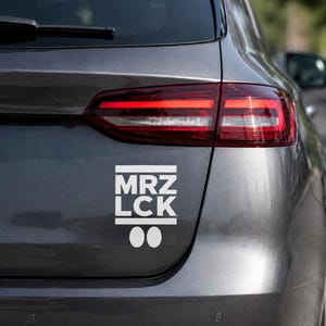 May include: A white sticker with the text "MRZ LCK" on the back of a gray car. The sticker has two white circles below the text. The car's taillight is visible.
