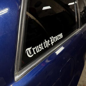 Puede incluir: Una puerta de coche azul con una calcomanía de vinilo blanco que dice "Trust the Process" en una fuente gótica. La calcomanía está aplicada a la ventana del coche. El coche está estacionado en el interior.