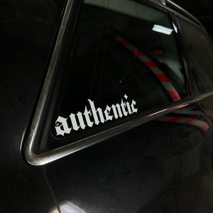Könnte beinhalten: Ein schwarzes Auto mit einem weißen "authentisch" Aufkleber auf der Heckscheibe.