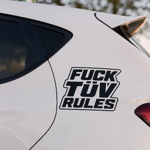 Könnte beinhalten: Ein weißes Auto zeigt einen schwarzen Aufkleber am hinteren Seitenteil. Der Sticker besteht aus fetten, blockartigen Buchstaben in drei Zeilen mit der Aufschrift FUCK TÜV RULES und einem dicken weißen Rand um den schwarzen Text auf dem Lack.