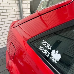 Könnte beinhalten: Ein rotes Auto mit einem weißen Aufkleber auf der Heckscheibe, auf dem "POLSKA AIRLINES" mit einem weißen Adlerlogo steht.