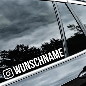 Op de afbeelding: Een witte vinyl sticker met een Instagram-icoon en de tekst "WUNSCHNAME" op een zwart autovenster.
