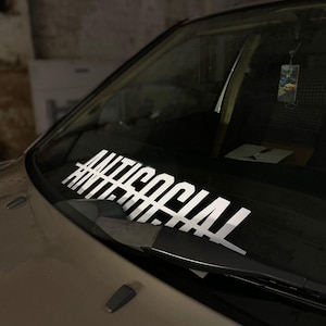 Könnte beinhalten: Nahaufnahme einer Windschutzscheibe eines Autos mit einem weißen Aufkleber, auf dem "ANTISOCIAL" steht.