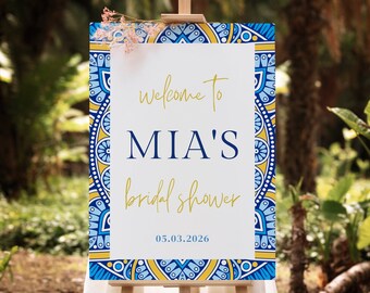 Op de afbeelding: Een wit bord met een blauwe en gele gepatroneerde rand. Het bord zegt "Welkom bij Mia's Bridal Shower 05.03.2026". Het bord staat op een ezel voor een groene struik.