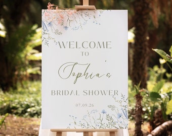 May include: A white sign with a floral border and the text "Welcome to Sophia's Bridal Shower 07.09.26". The sign is on an easel in a garden setting.