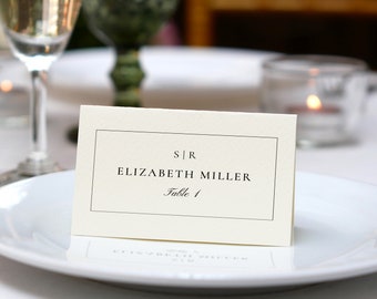 May include: A white place card with black text reads "S|R Elizabeth Miller Table 1" on a white tablecloth with a glass of champagne and a candle in the background.