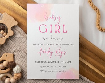 Puede incluir: Invitación a una fiesta de bienvenida al bebé de color rosa y blanco con fondo de acuarela. El texto dice "baby GIRL is on her way". La invitación incluye el nombre "Hailey Keys", la fecha "sábado 25 de noviembre a las 5:00 PM", la dirección "123 Anywhere St., Any City" y la información de RSVP.