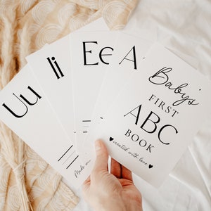 May include: A set of white rectangular cards featuring black printed letters and text. The top card displays the title Baby's FIRST ABC BOOK with the phrase created with love and two small heart icons. Other cards show letters Uu, Ii, Ee, and Aa.