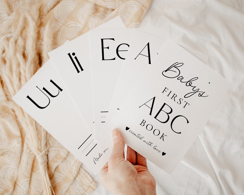 May include: A set of white rectangular cards featuring black printed letters and text. The top card displays the title Baby's FIRST ABC BOOK with the phrase created with love and two small heart icons. Other cards show letters Uu, Ii, Ee, and Aa.