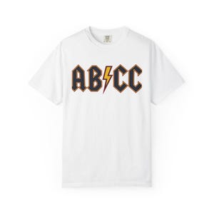 May include: A white short-sleeve t-shirt featuring the text AB/CC printed across the chest. The letters are dark navy blue with a thin orange outline. A yellow lightning bolt with a thin orange outline separates the letters B and C.