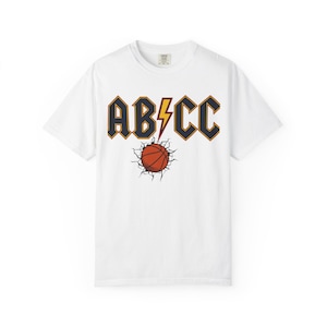 May include: A white short-sleeve t-shirt featuring a graphic print on the chest. The design displays the text ABCC in dark blue with orange outlines, a yellow lightning bolt, and an orange basketball breaking through a cracked surface.
